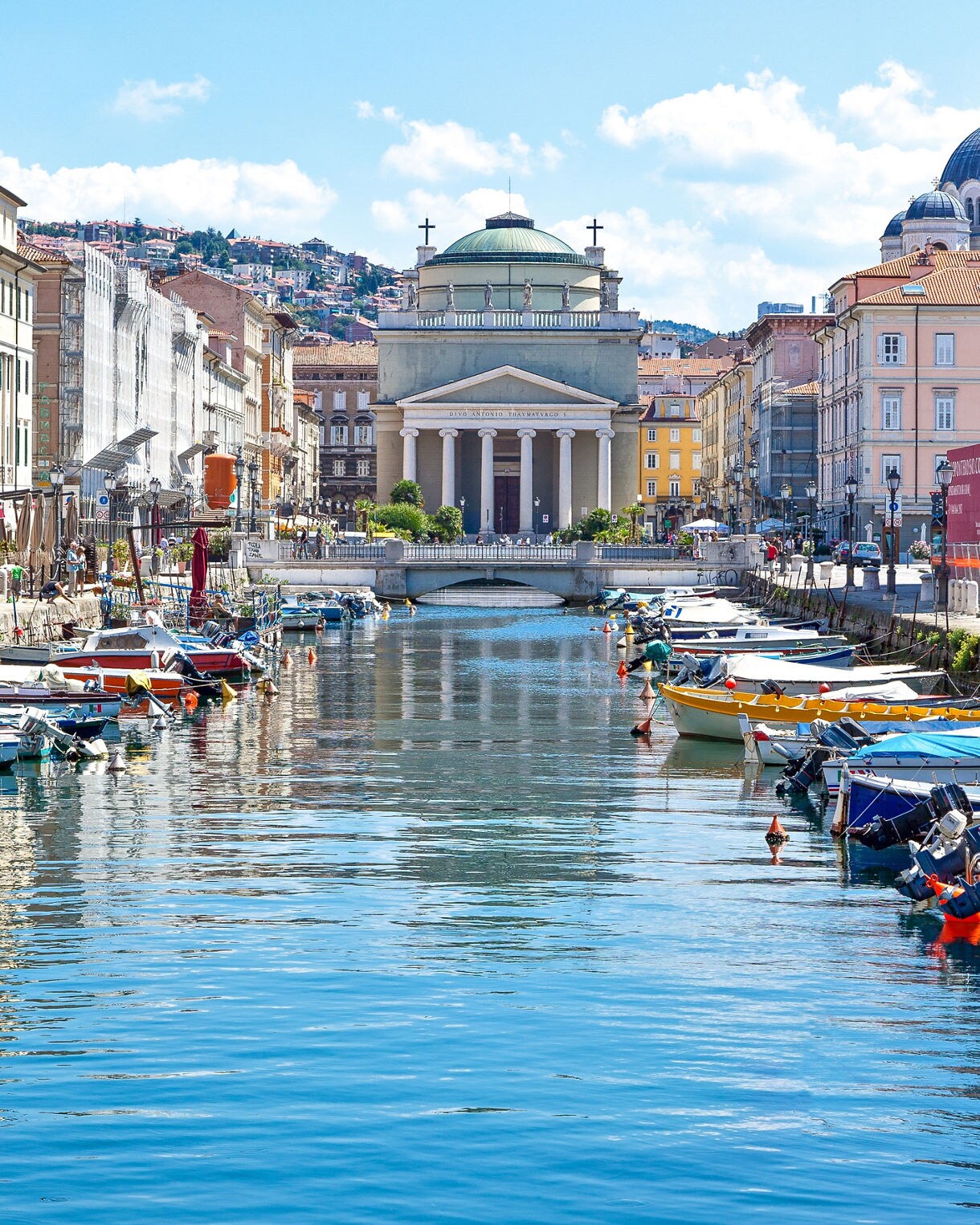 Vertical view of Trieste’s Canal Grande lined with boats and colorful buildings, leading toward the Neoclassical Sant’Antonio Taumaturgo church under a bright blue sky.