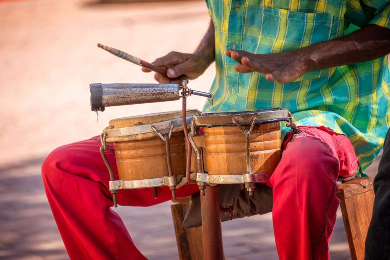 A street musician in Trinidad, Cuba, playing traditional drums while wearing a bright checkered shirt and red pants, creating rhythmic beats in the vibrant atmosphere of the city.