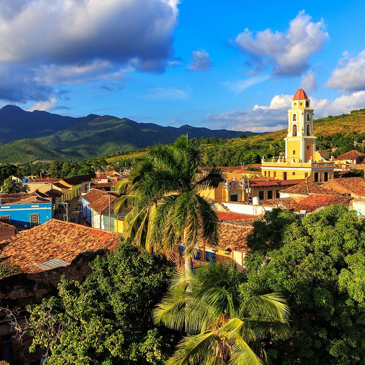 Aerial view of historic town with terracotta roofs, church bell tower, palm trees, and green mountains in background.