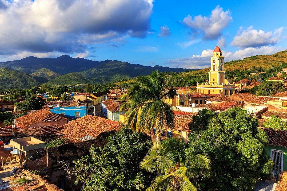 A scenic view of Trinidad, Cuba, showcasing red-tiled rooftops, a colonial bell tower, lush greenery and the Escambray Mountains in the background.