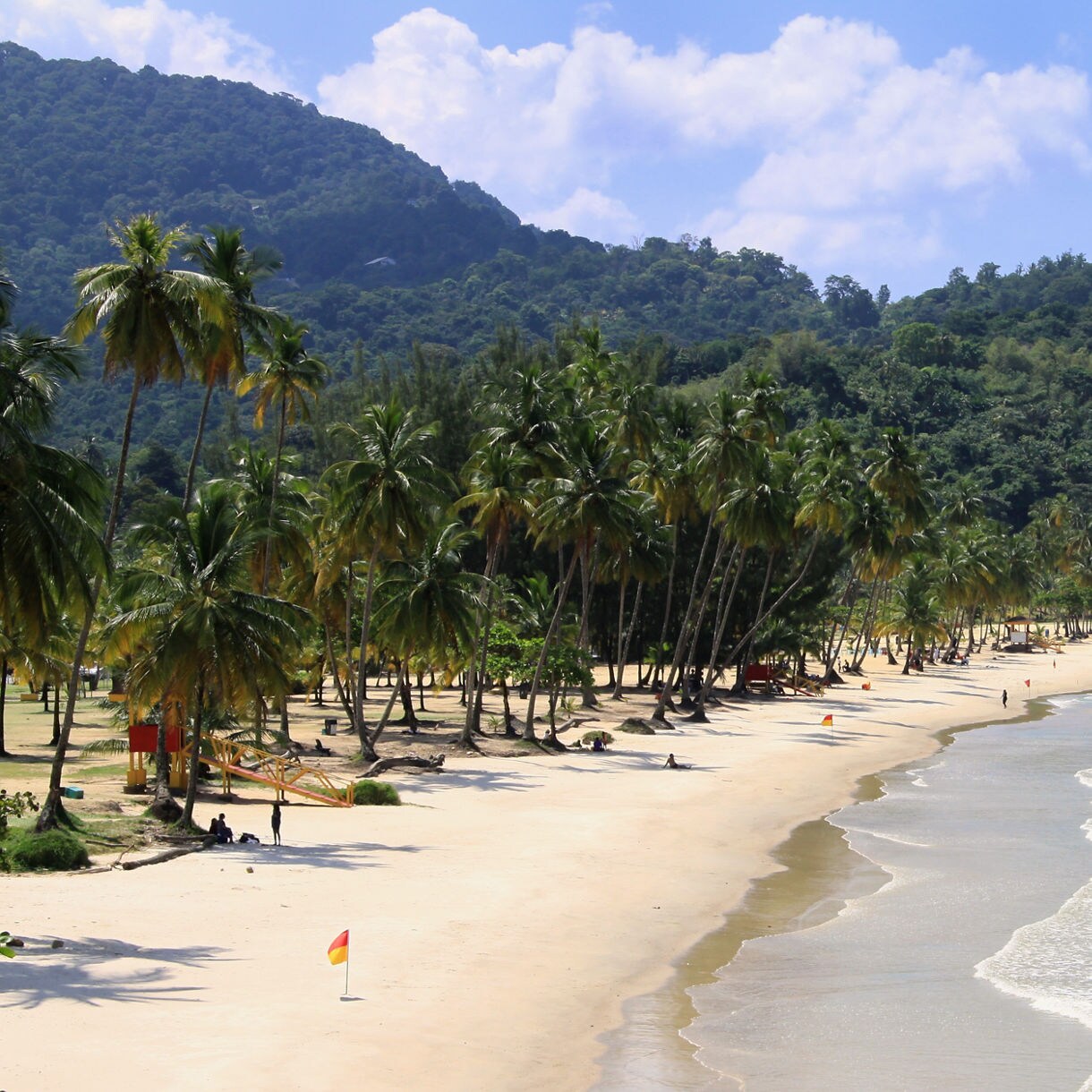 Curved beach with white sand bordered by dense green forest and hills under partly cloudy sky.