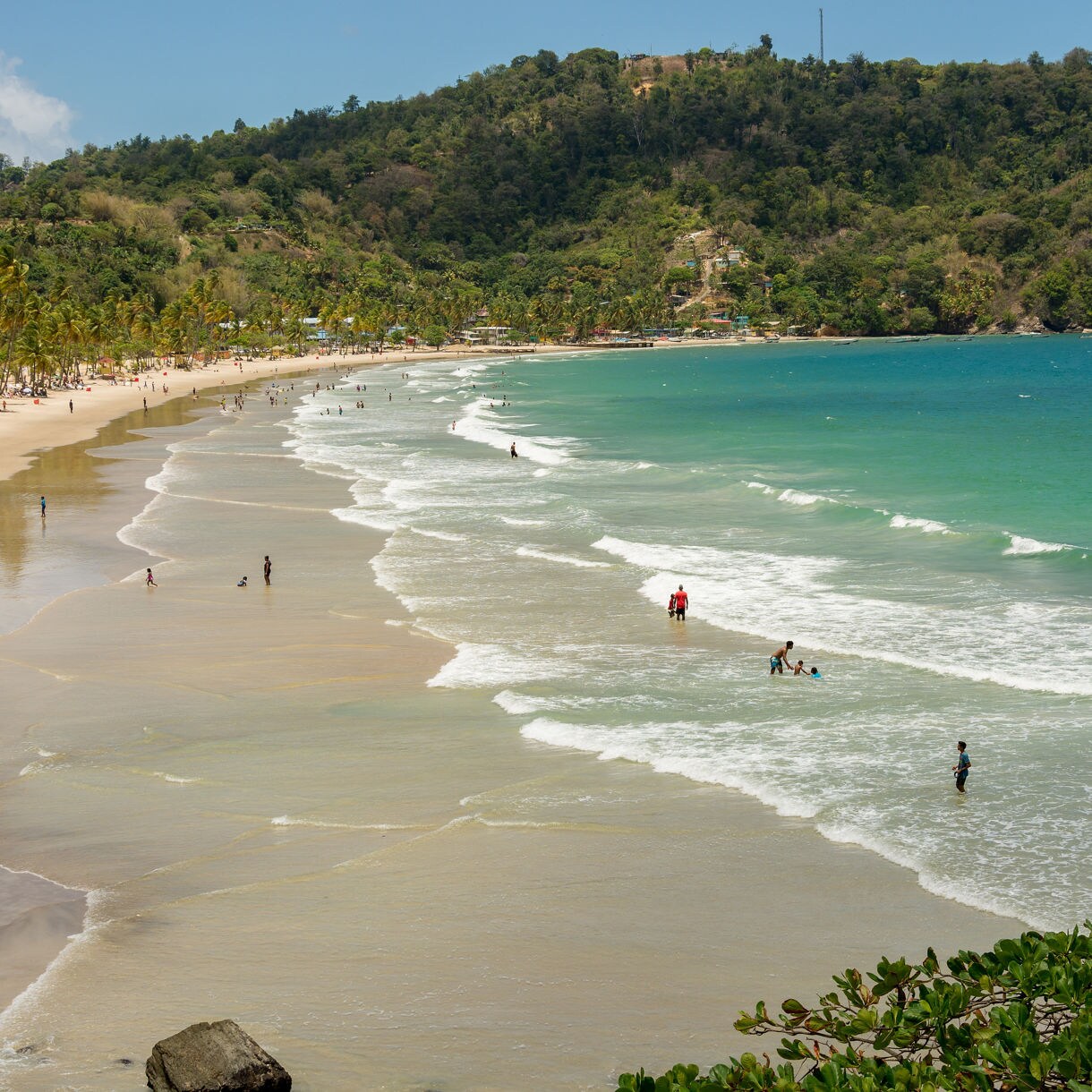 A scenic view of Maracas Beach in Trinidad, with gentle waves lapping the shore and visitors enjoying the water, backed by green hills.