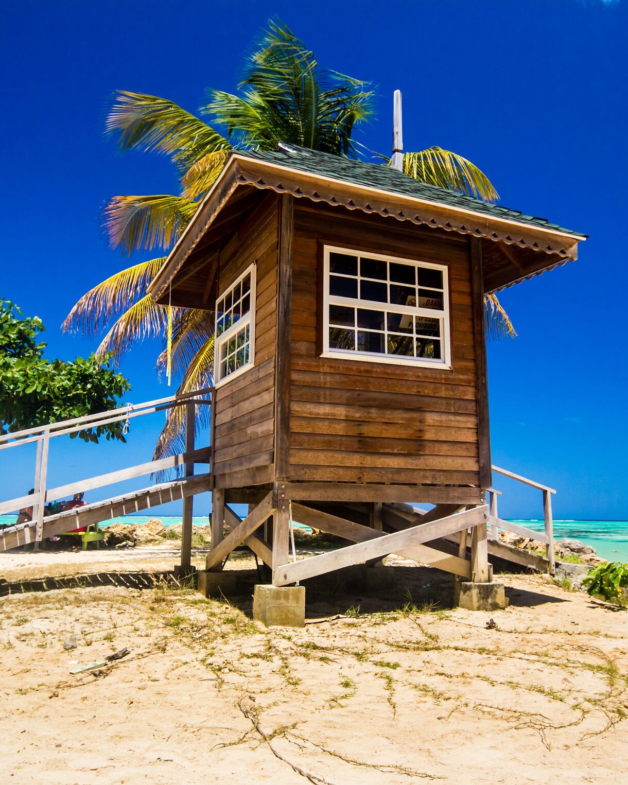 A wooden lifeguard hut on stilts at Pigeon Point Beach, Trinidad, with a backdrop of clear turquoise waters, white sand and lush palm trees.