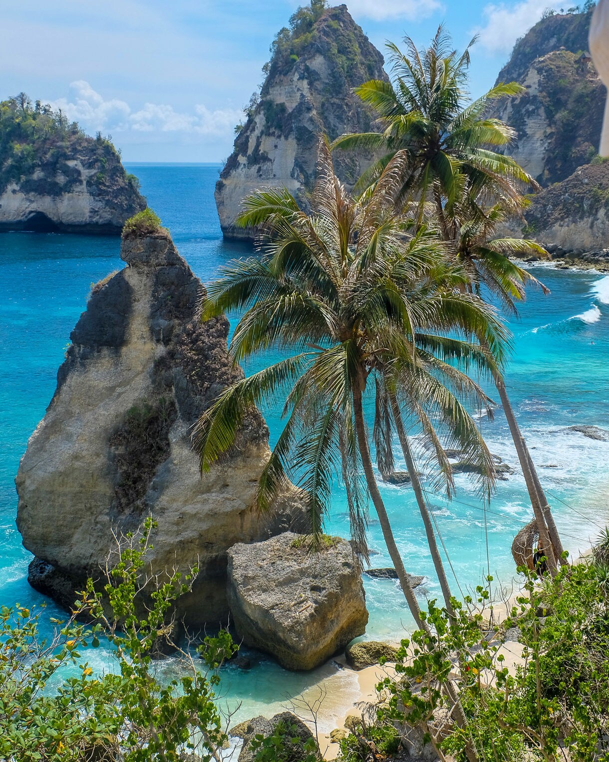 Tropical coastal scene with tall limestone rock formations, turquoise waves, leaning palm trees and a small sandy cove surrounded by lush greenery.