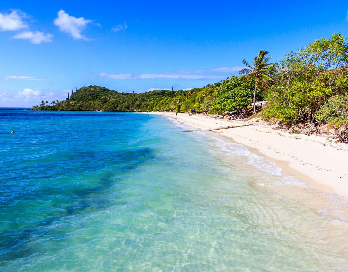 White sand beach with clear turquoise water, gentle waves and lush green trees lining the coast under a bright blue sky.