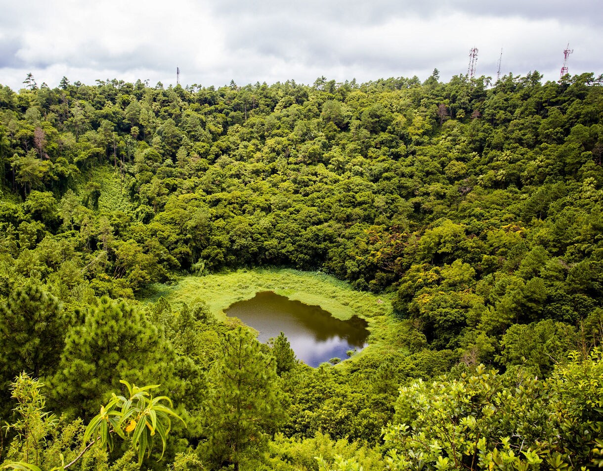 A small pond sits in the center of a densely forested volcanic crater, surrounded by thick green trees.