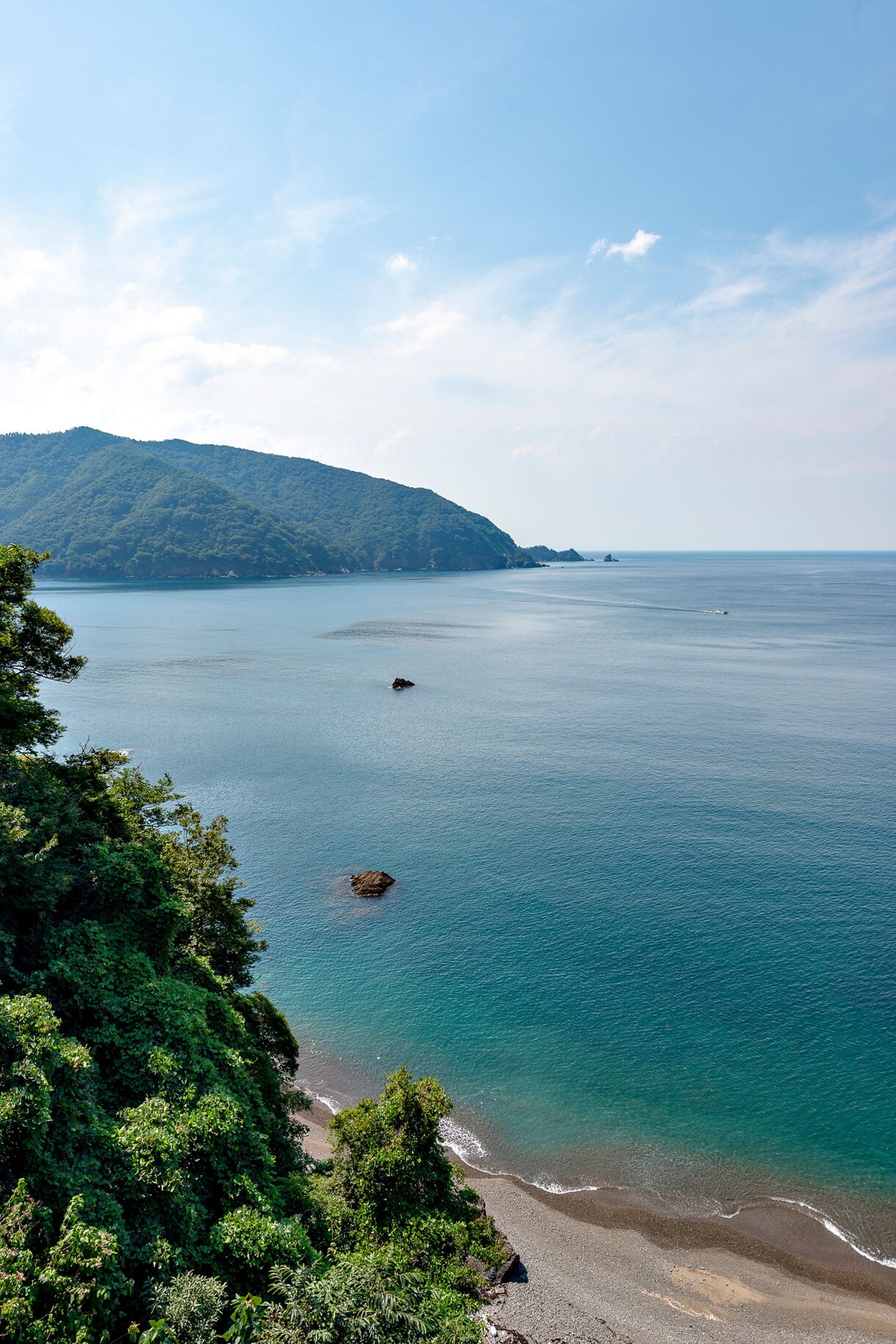 View of Tsuruga’s coastline with calm blue water, forested hills and rocky shoreline under a clear sky.