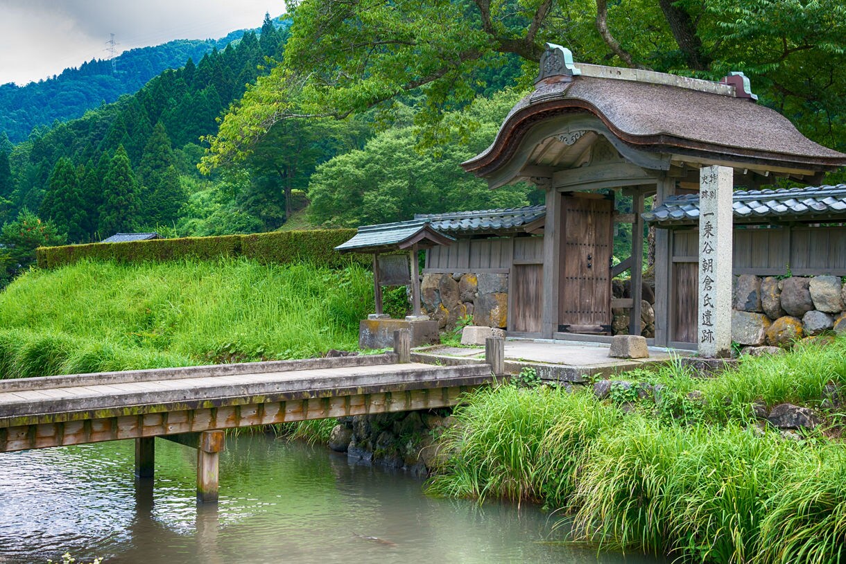 Historic wooden gate and footbridge at Ichijōdani Asakura Clan Ruins surrounded by tall grass, stone walls and forested hills.