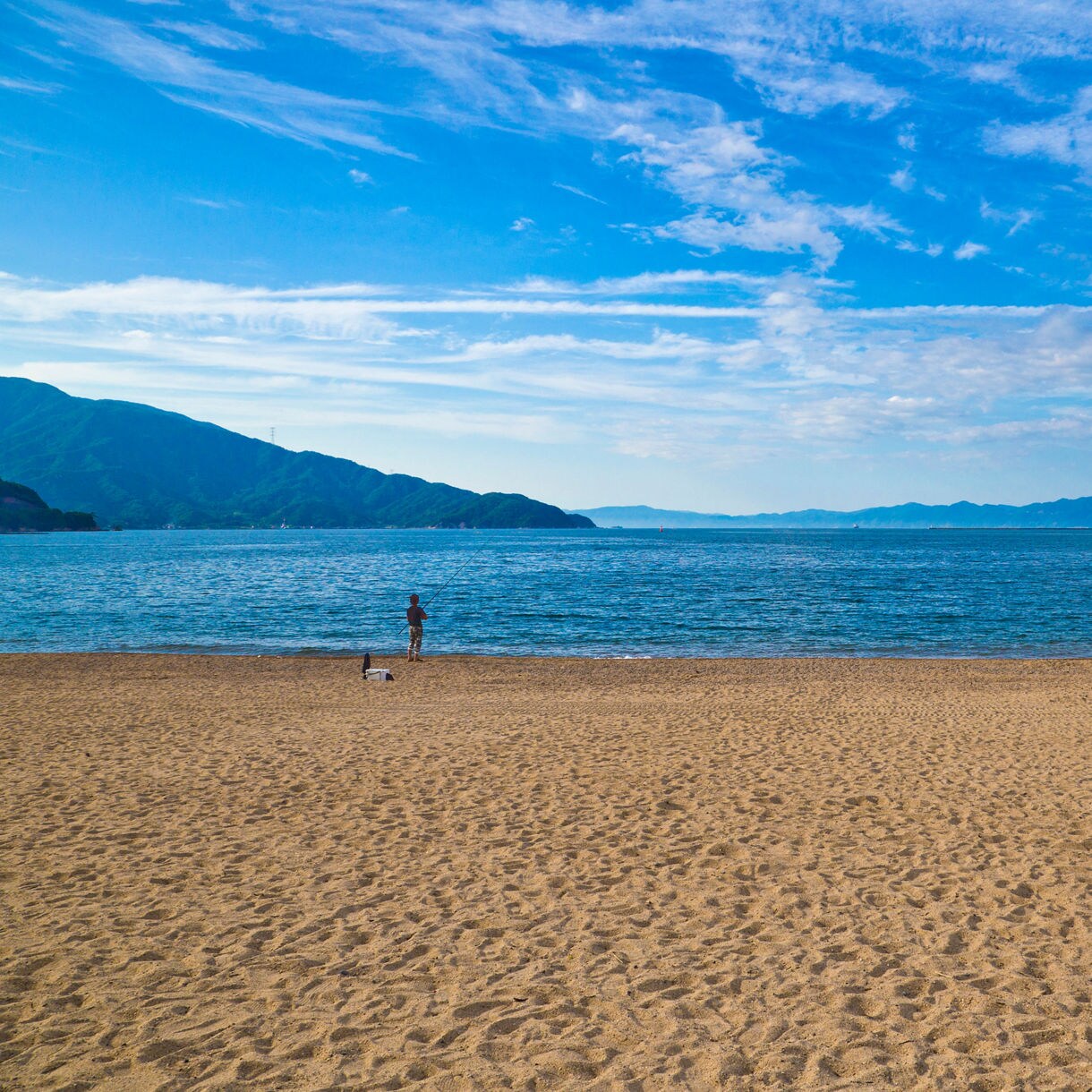 Expansive sandy beach at Kehi-no-Matsubara with a lone fisherman standing by the shoreline, calm deep-blue water and distant forested mountains beneath a bright sky with scattered clouds.