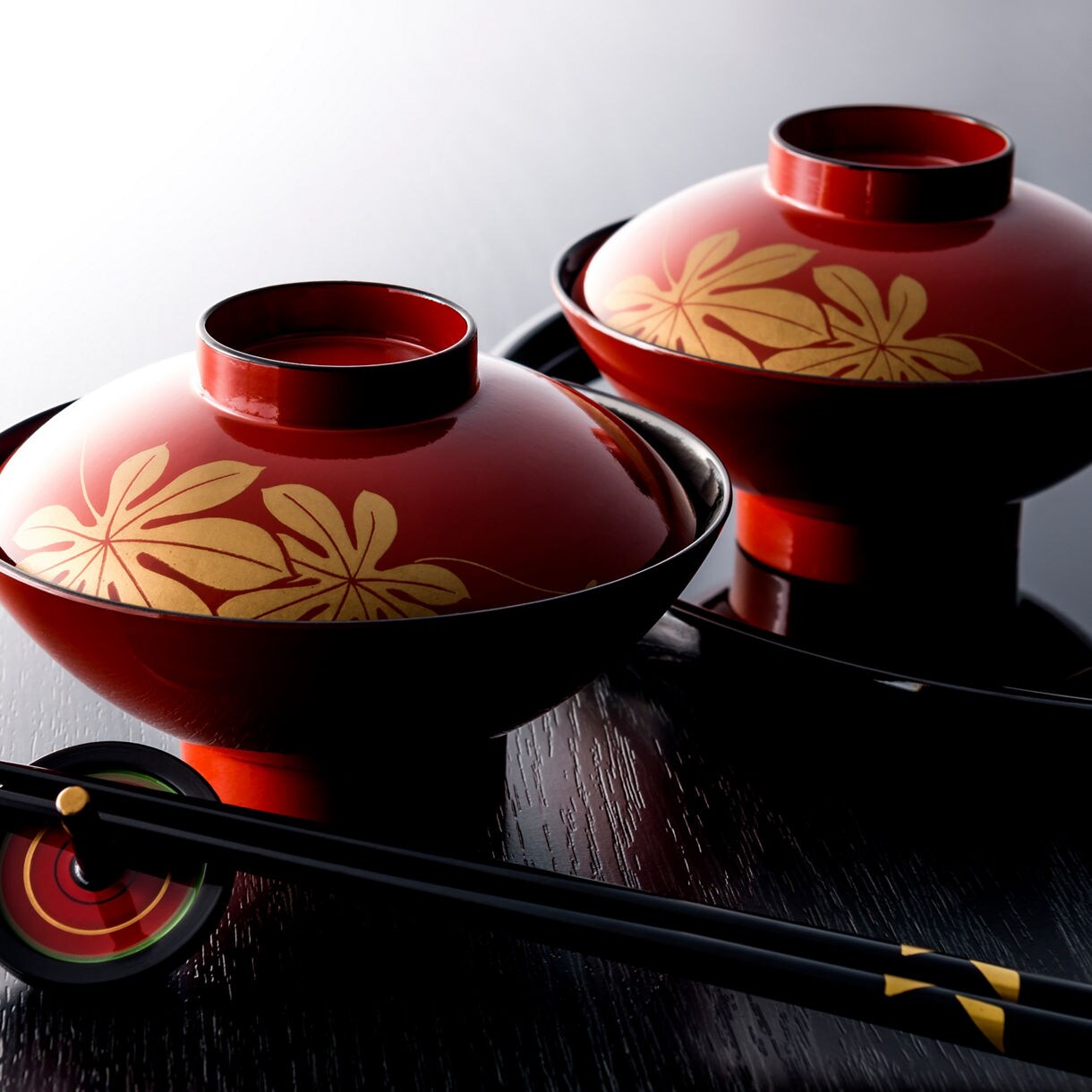 Two red lacquered bowls with gold leaf designs and black chopsticks resting on a glossy black surface.