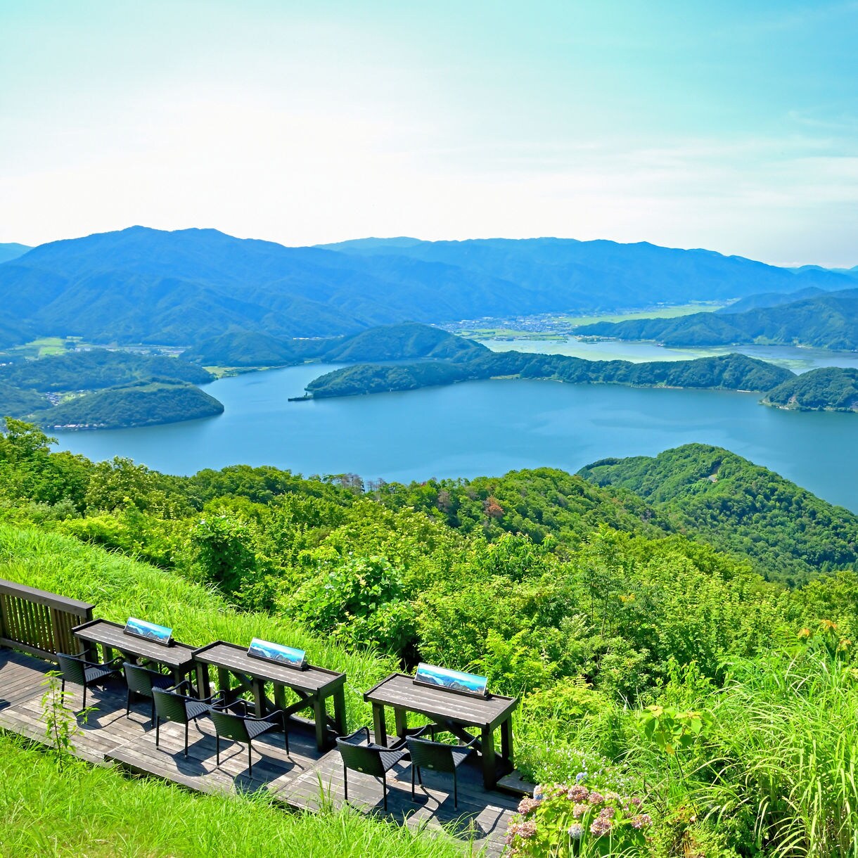Scenic view from a hilltop terrace overlooking Mikata Five Lakes, surrounded by forested mountains under a bright blue sky. Tables and chairs are set along a wooden deck in the foreground.