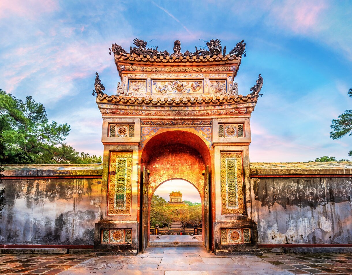 Ornate entrance gate at Tu Duc’s Royal Tomb in Hue, Vietnam, featuring colorful carvings, tiled roofs and a sunlit path leading to ancient pavilions beyond.