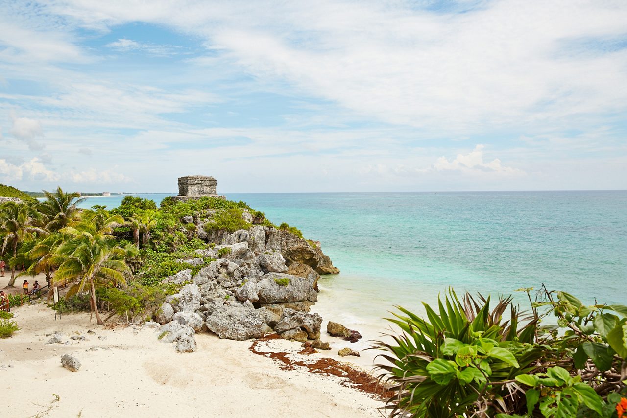 View of the ancient Mayan ruins in Tulum, Mexico, overlooking a pristine beach and turquoise waters, surrounded by lush tropical vegetation.