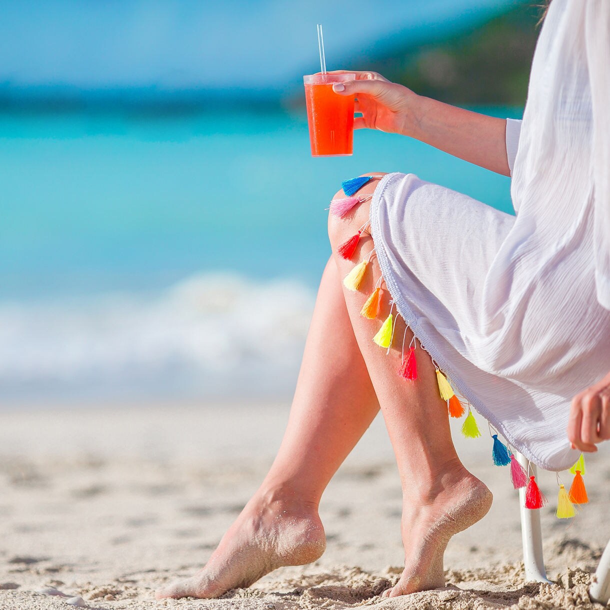 A person enjoying a refreshing drink on the beach, sitting in the soft sand near the crystal-clear water, with colorful tassels decorating their beach attire.