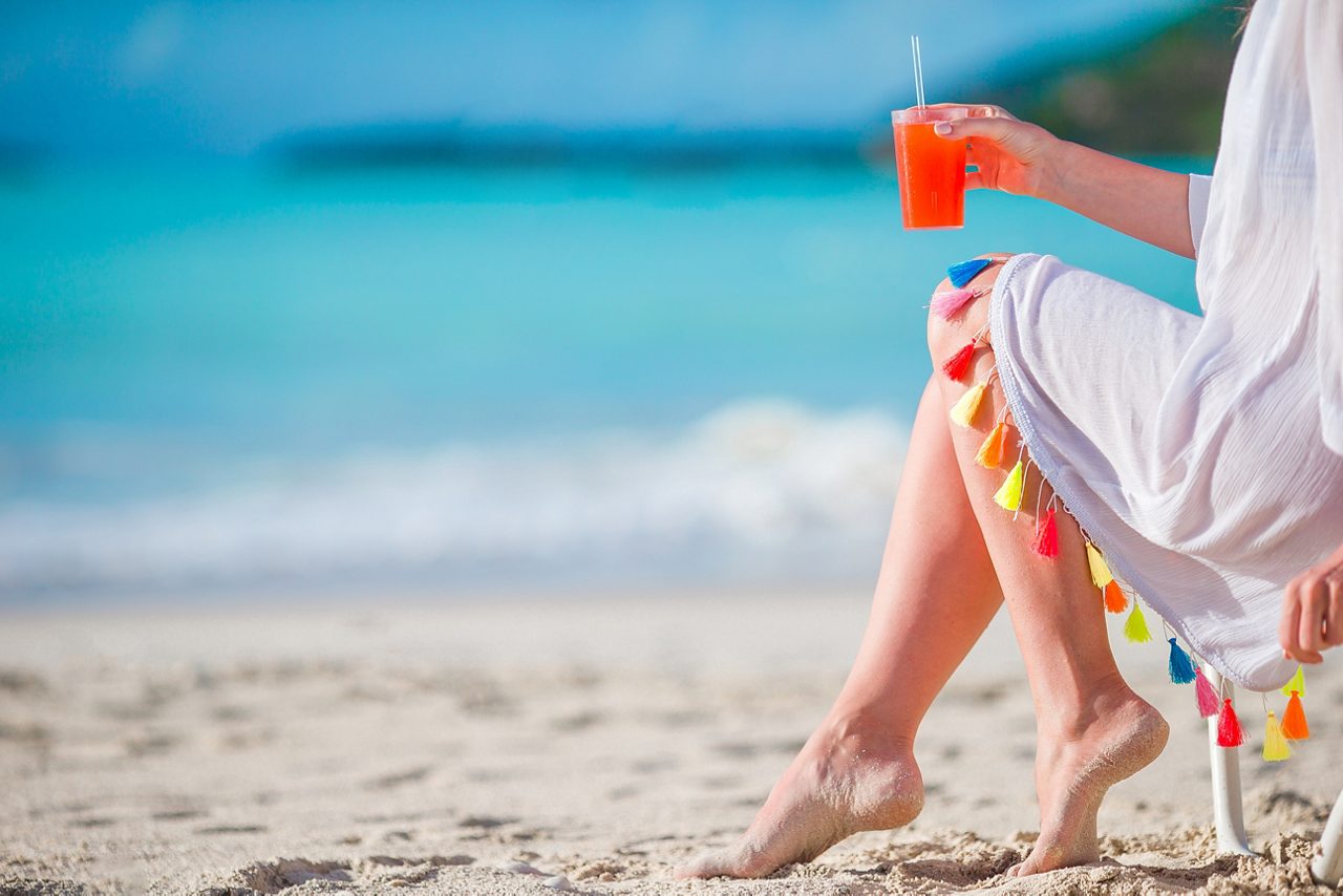 A person relaxing on a beach chair with their feet in the sand, holding a red tropical drink, with clear blue ocean water and sky in the background