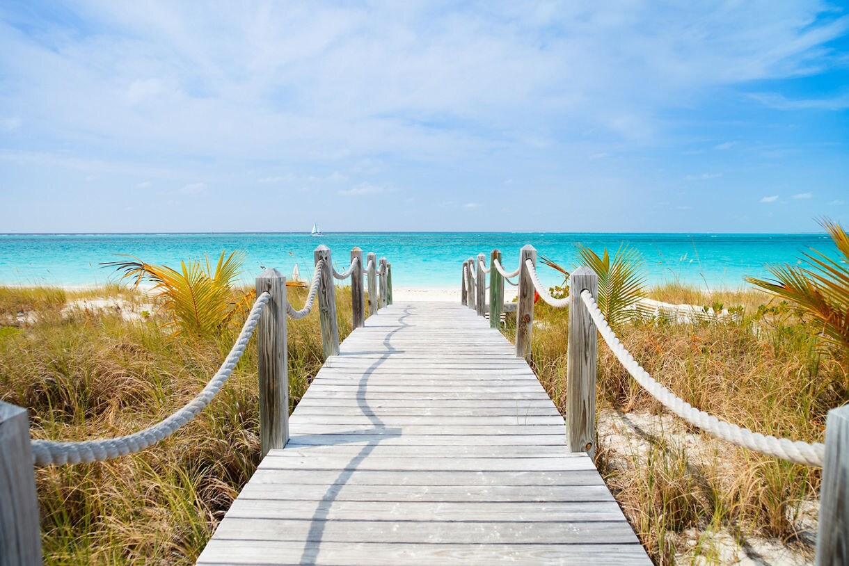 A weathered wooden boardwalk with rope railings leading to a white sand beach in Turks and Caicos, surrounded by coastal grass and palm fronds.
