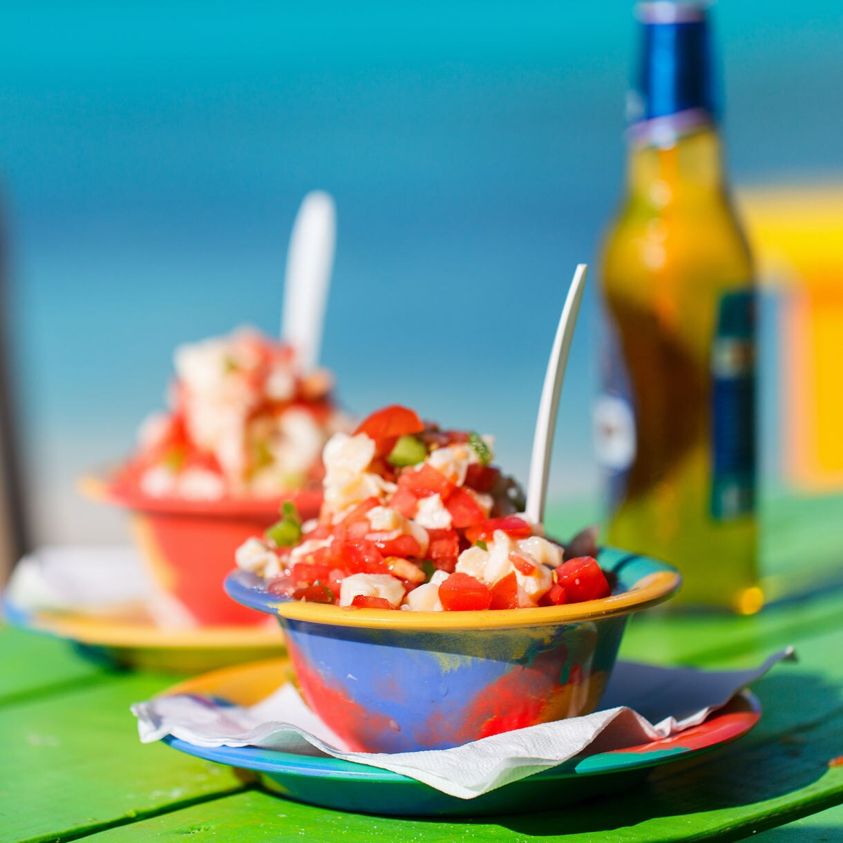 Two vibrant bowls of conch salad on a colorful table, with fresh tomatoes, onions and conch meat, set against the background of a turquoise beach and a cold drink nearby.