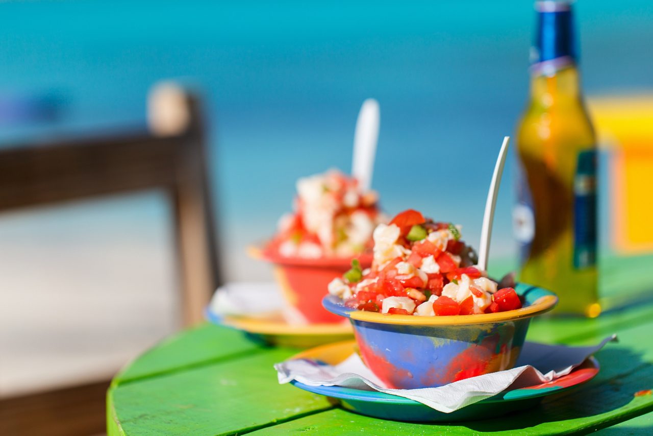 Two vibrant bowls of conch salad on a colorful table, with fresh tomatoes, onions and conch meat, set against the background of a turquoise beach and a cold drink nearby.