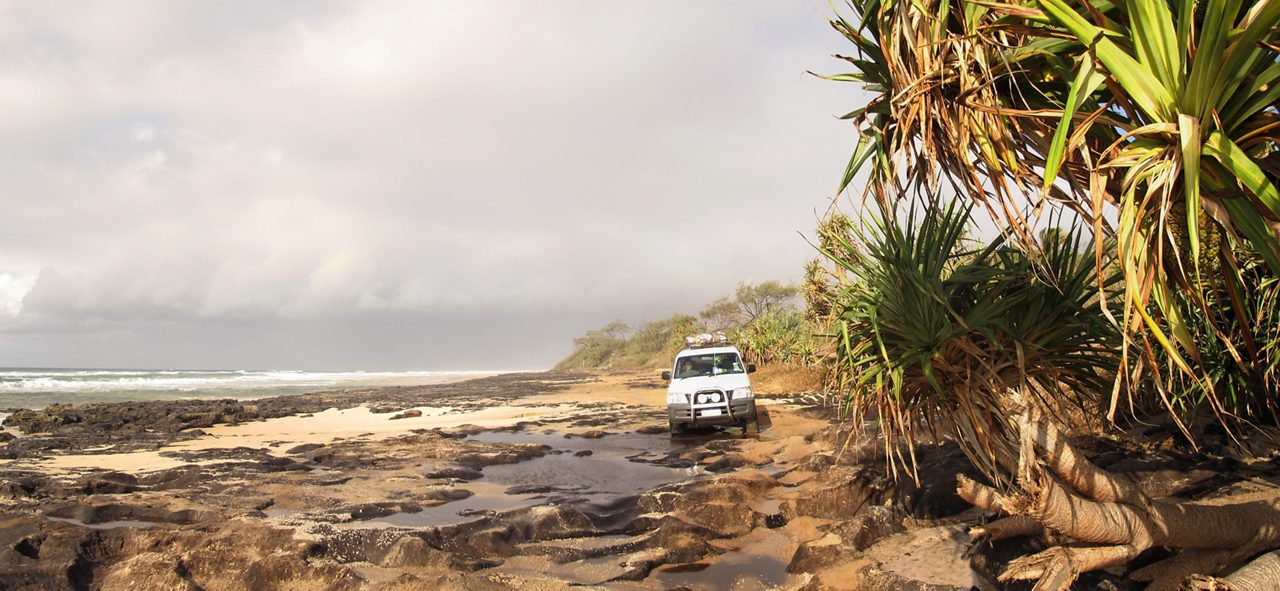 A Jeep driving along a rugged beach path surrounded by lush vegetation and a vast ocean view in the background.