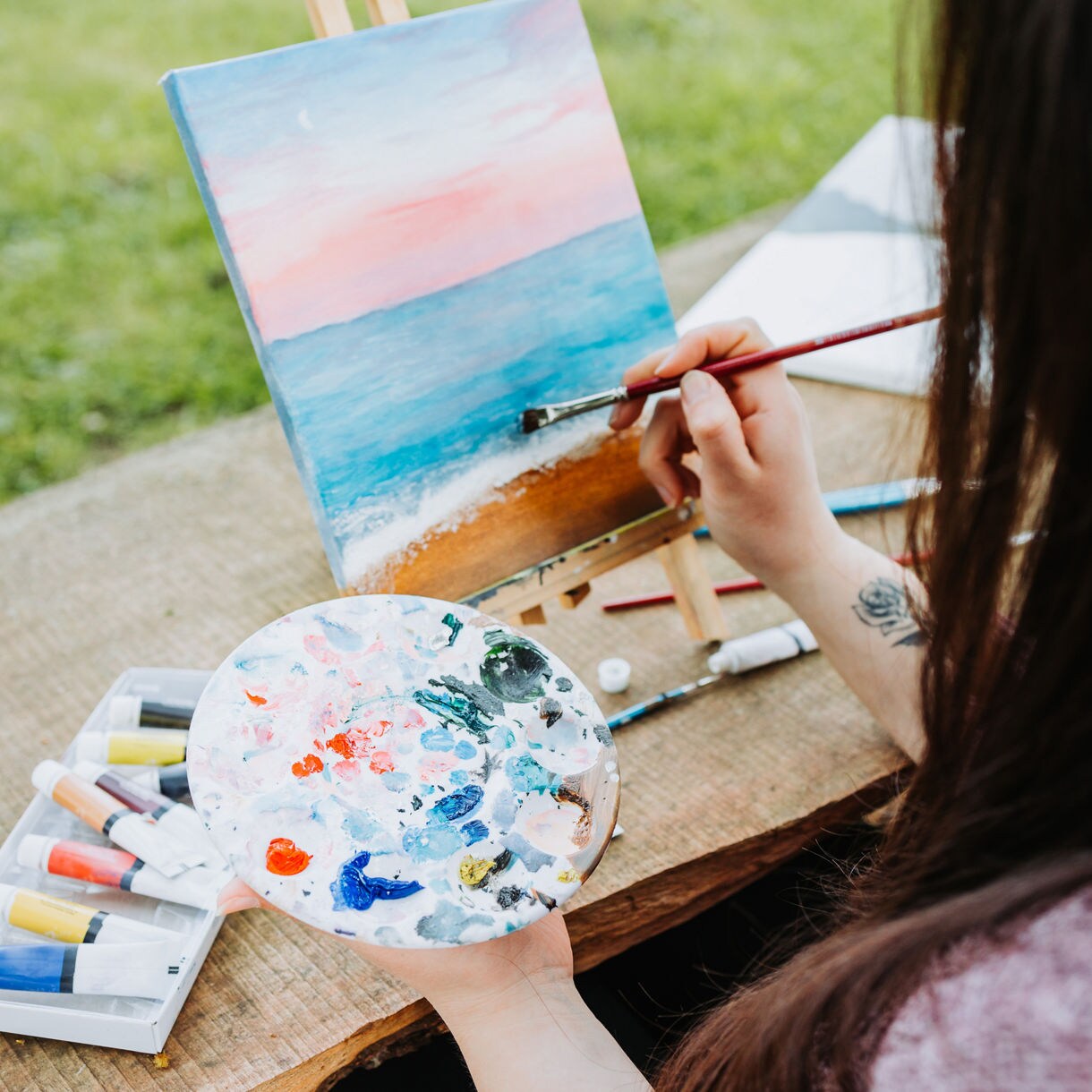 A person painting a tranquil beach scene on a canvas, holding a palette with vibrant paint colors, as the island's lush greenery and soft sky provide inspiration.