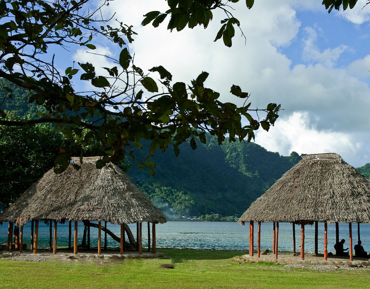 Two traditional thatched-roof Samoan fales sit on a grassy shoreline beside a calm blue bay, with people sitting inside one and lush green mountains rising in the background.