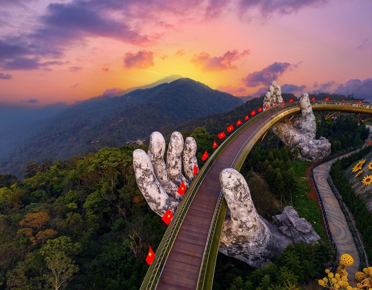 The Golden Bridge in Ba Na Hills, Da Nang, Vietnam, at sunset, featuring two massive stone hands supporting a curved walkway lined with red flags above a lush forest.