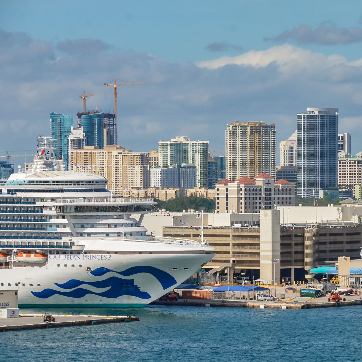 Two large cruise ships are docked at a bustling urban port with a city skyline in the background. The ships feature distinctive wave-like designs on their hulls. The setting includes high-rise buildings, cranes, and a clear blue sky, suggesting a vibrant coastal city environment.