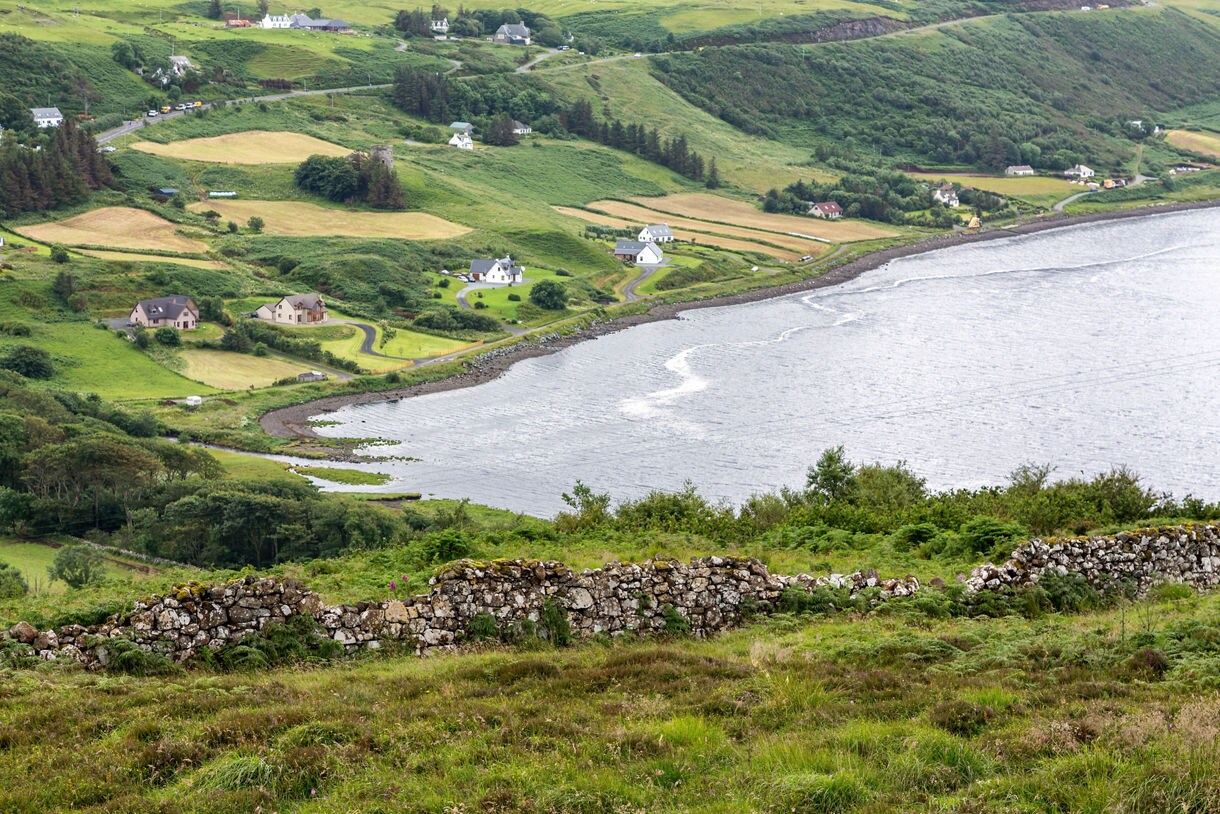 Elevated view of Uig Bay on the Isle of Skye, showing patchwork fields, scattered cottages, winding roads and a curving shoreline backed by lush green hills.