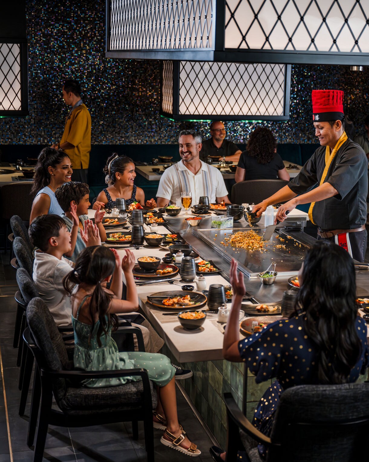 A teppanyaki chef performs cooking at a hibachi grill while guests are seated around the cooking surface in a modern Japanese restaurant with decorative lighting fixtures. 