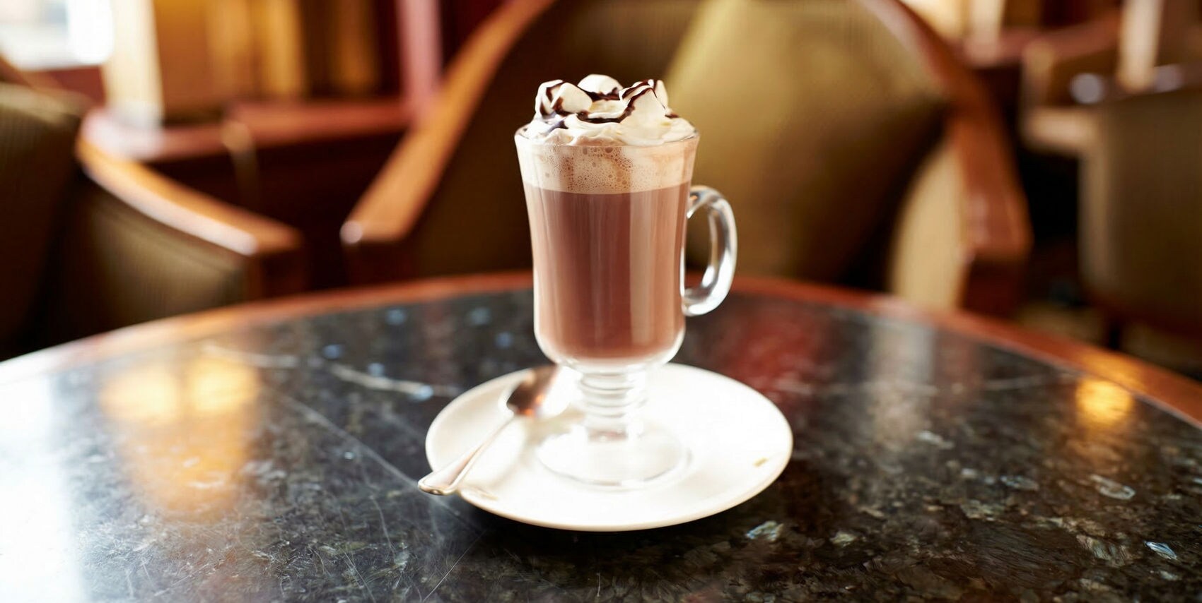 A glass mug filled with hot chocolate topped with whipped cream and chocolate drizzle sits on a white saucer with a spoon. The drink is placed on a marble table in a cozy café setting with upholstered chairs in the background. Warm lighting and soft brown tones create a relaxed and inviting atmosphere. No visible text or numbers are present in the image.