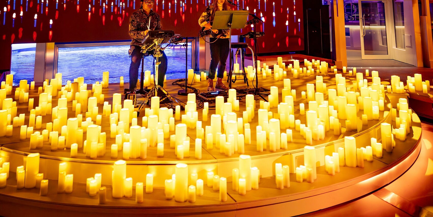 Two musicians are seated and performing on a circular stage filled with numerous glowing candles. The setting features a dramatic arched ceiling with reflective surfaces and warm, golden lighting. The background includes a digital display of hanging lights, enhancing the immersive ambiance. The overall mood is intimate and atmospheric, with a focus on live music in a visually striking environment.