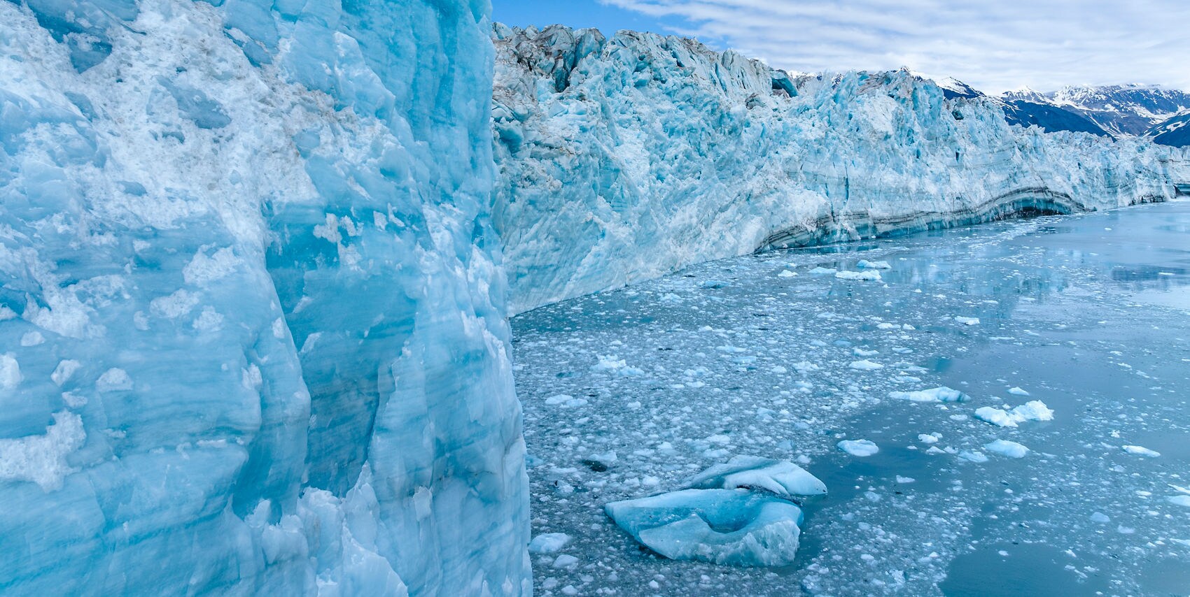 A towering blue glacier dominates the left side of the image, with jagged ice formations stretching into the distance. The scene is set in a cold, remote environment, with floating ice chunks scattered across the calm water. Snow-capped mountains are visible in the background under a partly cloudy sky, emphasizing the frigid and pristine atmosphere.