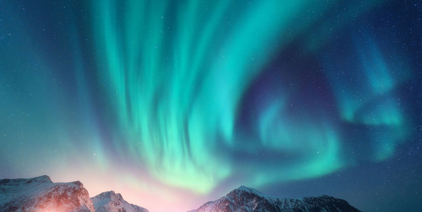 A striking display of green and blue northern lights illuminates the night sky above snow-covered mountains. The aurora reflects beautifully on the calm water below, creating a surreal and tranquil atmosphere. The scene is set in a remote, natural environment with no visible people or text. The overall mood is serene and awe-inspiring, with cool tones dominating the palette.