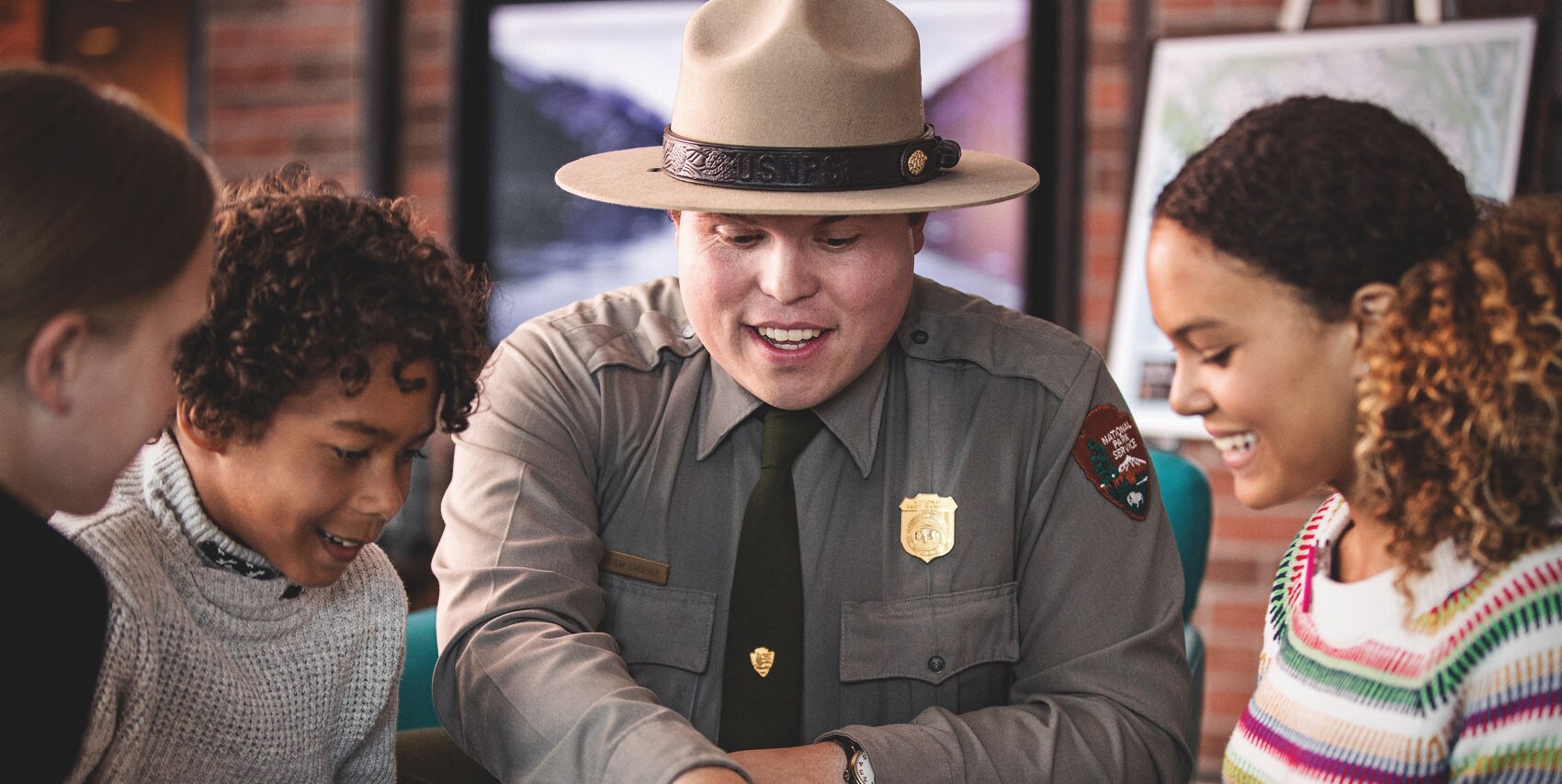 A park ranger in uniform sits at a table with a group of children, engaging them in an educational activity. The setting appears to be indoors, possibly a visitor center or classroom, with nature-themed visuals in the background. The ranger points to an object on the table, encouraging curiosity and learning. The children are of diverse backgrounds and appear attentive and interested.