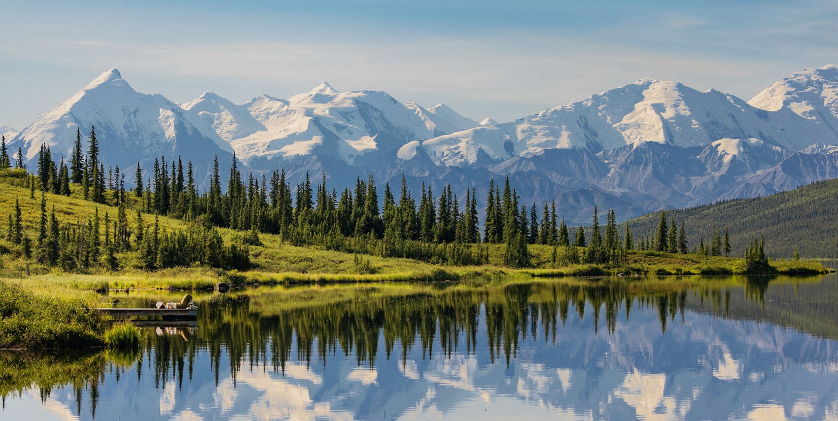 A serene lake mirrors a range of snow-capped mountains under a clear blue sky. Evergreen trees line the shore, with lush green grass adding vibrant color to the scene. The tranquil water creates a perfect reflection of the dramatic peaks and forest. A small boat with two people is visible near the shore, enhancing the peaceful, natural setting. The overall mood is calm and picturesque, with crisp, natural light.