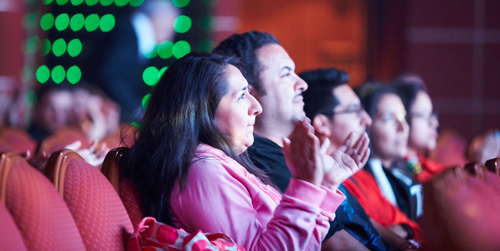 A group of people is seated in a theater, clapping and watching an event on stage. The setting features red cushioned chairs and a softly lit ambiance with green lights in the background. The audience members are dressed casually, suggesting a relaxed and informal atmosphere. The image captures the engagement and appreciation of the crowd during a live performance.
