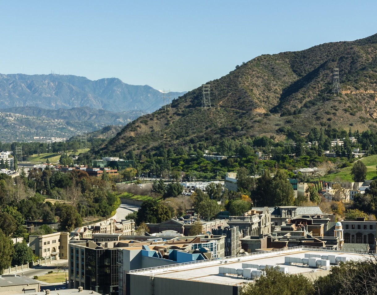 A wide elevated view of Universal Studios’ backlot with clustered soundstages, faux city facades and green hills rising behind them under a clear sky.