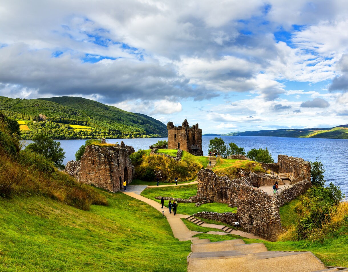 Ruins of Urquhart Castle on a grassy hilltop beside Loch Ness, with rolling green hills and dramatic clouds in the background.