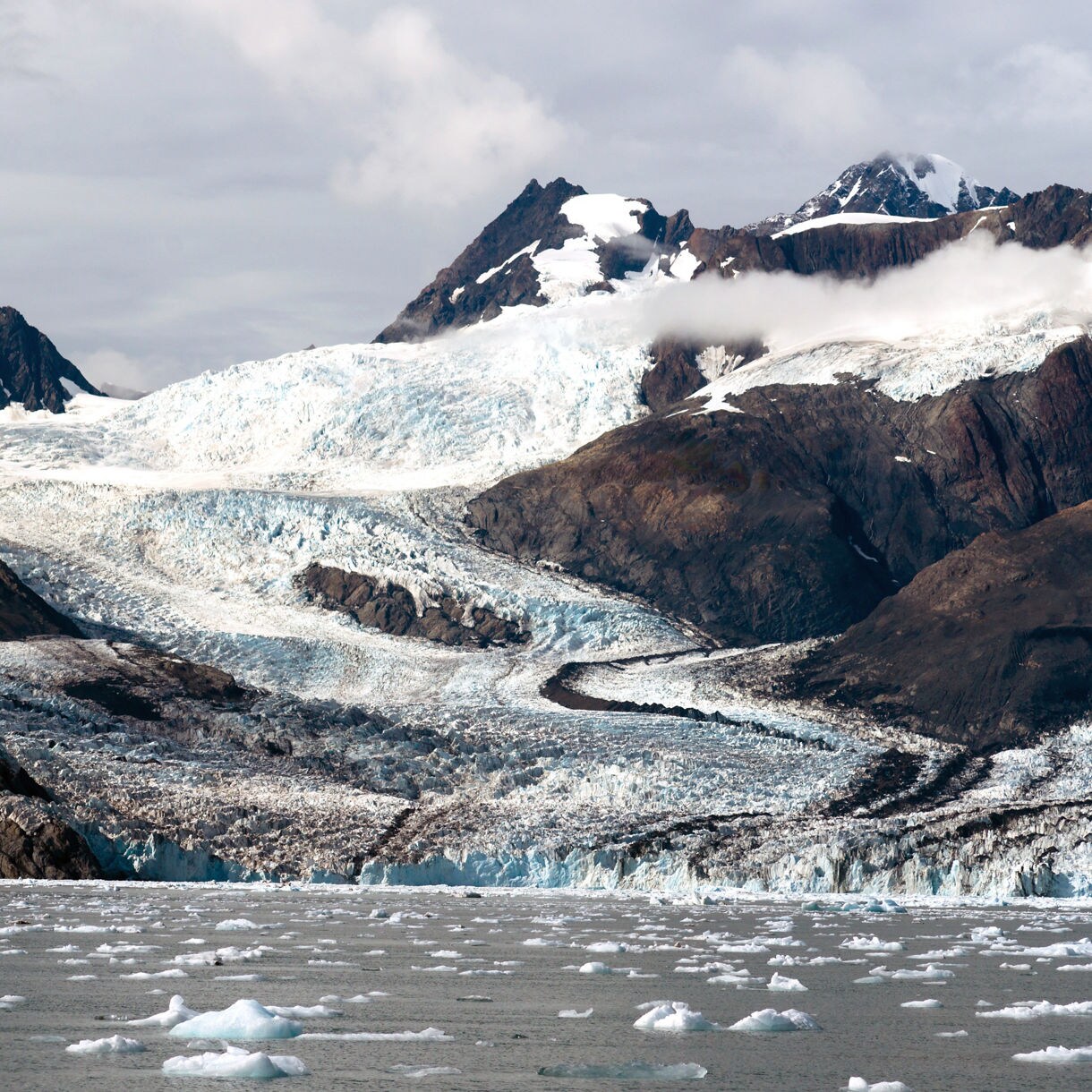 Massive glacier flowing between dark, rocky mountains near Valdez, Alaska with chunks of ice floating in the cold gray water below.
