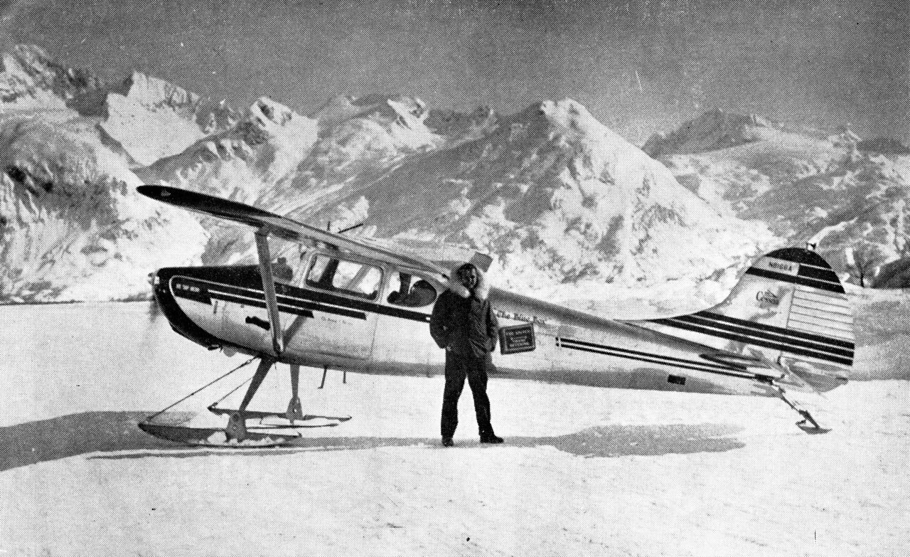 A black-and-white photograph of Bishop Gordon and the "Flying Blue Box" at Valdez, Alaska, with snow-capped mountains in the background.