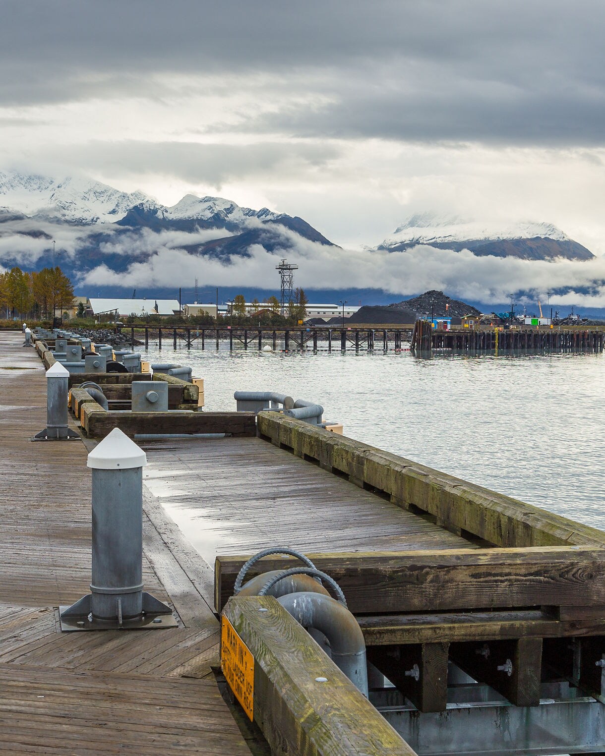 View of the wooden dock and port area in Valdez, Alaska on a cloudy day, with snow-capped mountains and low-hanging clouds in the background.