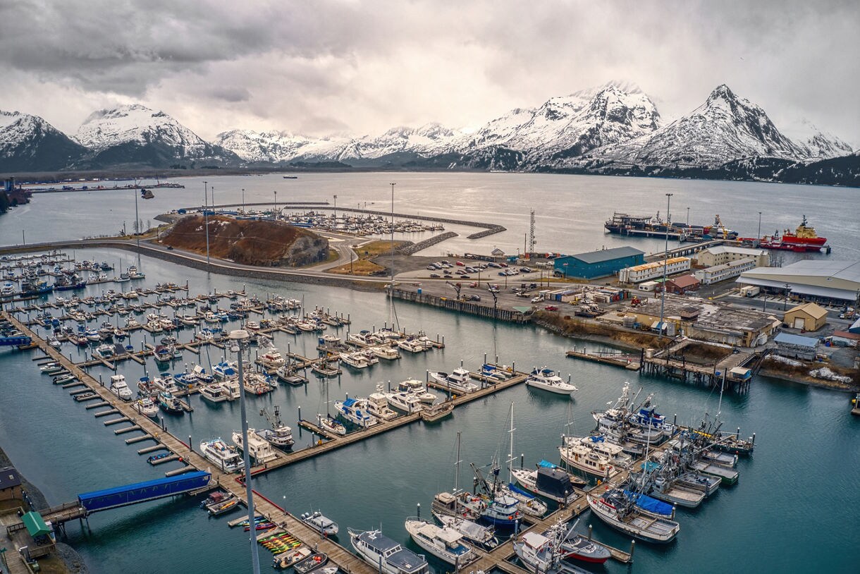 Aerial view of Valdez, Alaska, in spring, featuring a marina filled with boats, industrial docks, and snow-covered mountains in the background under a cloudy sky.