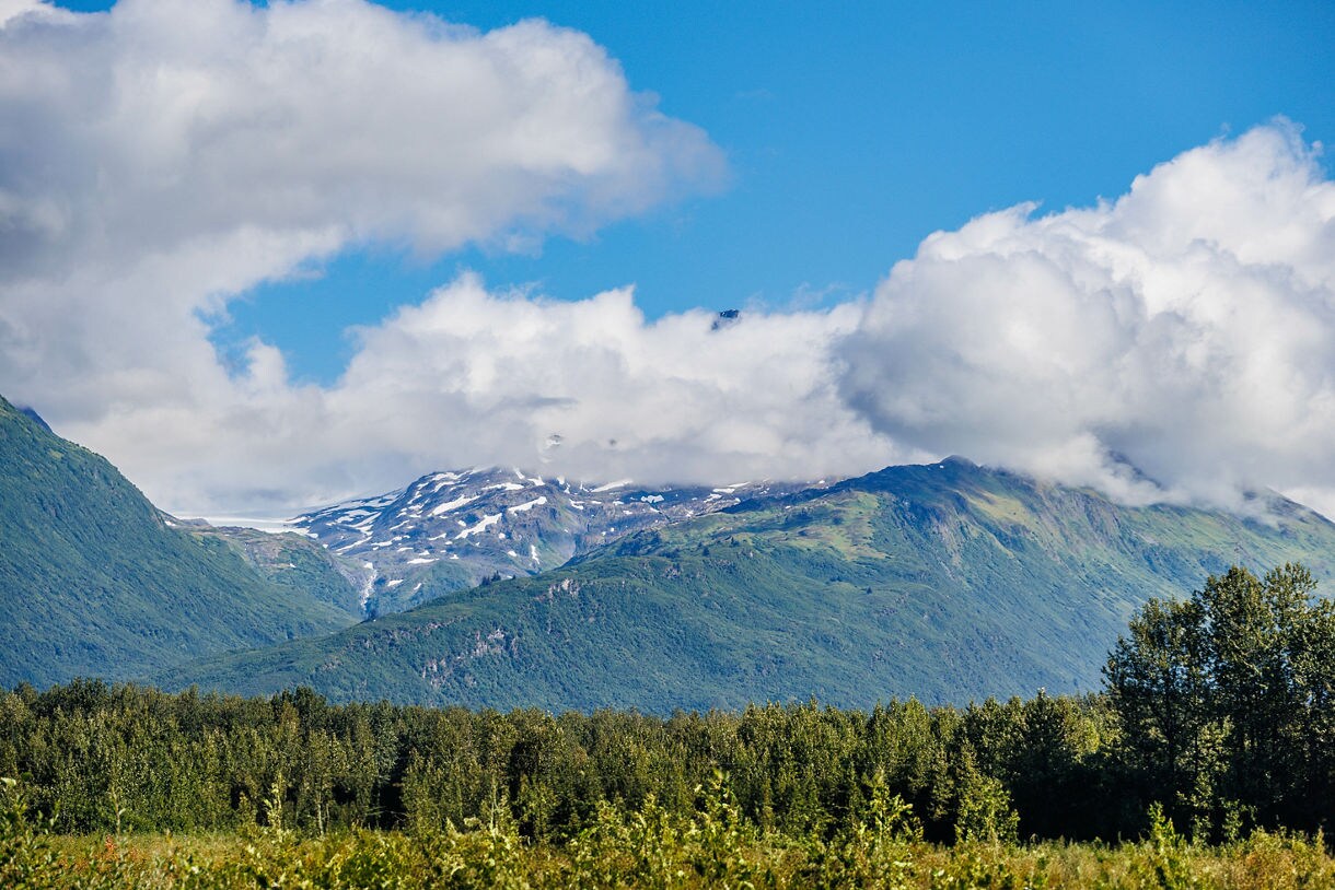 View of Thompson Pass near Valdez, Alaska with forested hills in the foreground, green mountains streaked with snow and low-hanging clouds beneath a bright blue sky.