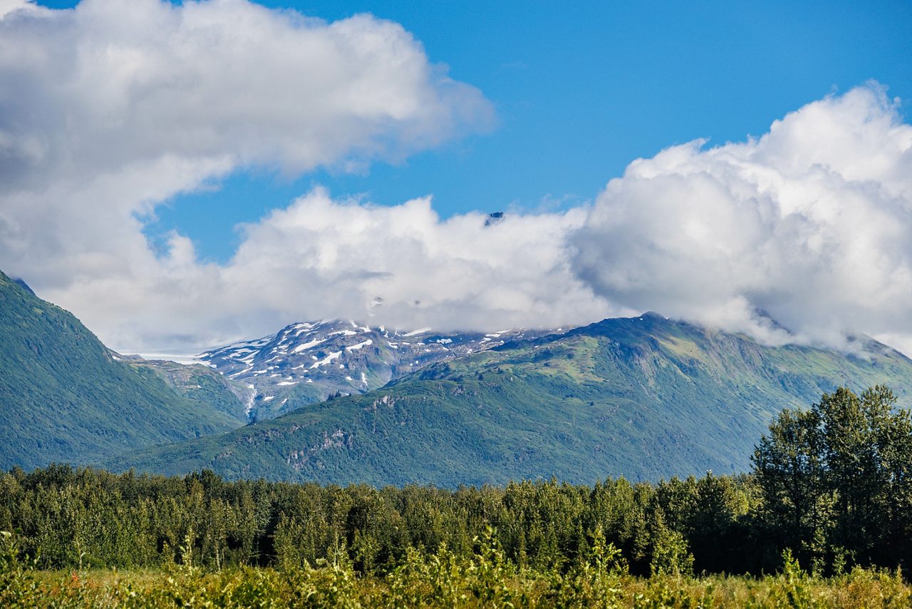 View of Thompson Pass near Valdez, Alaska with forested hills in the foreground, green mountains streaked with snow and low-hanging clouds beneath a bright blue sky.
