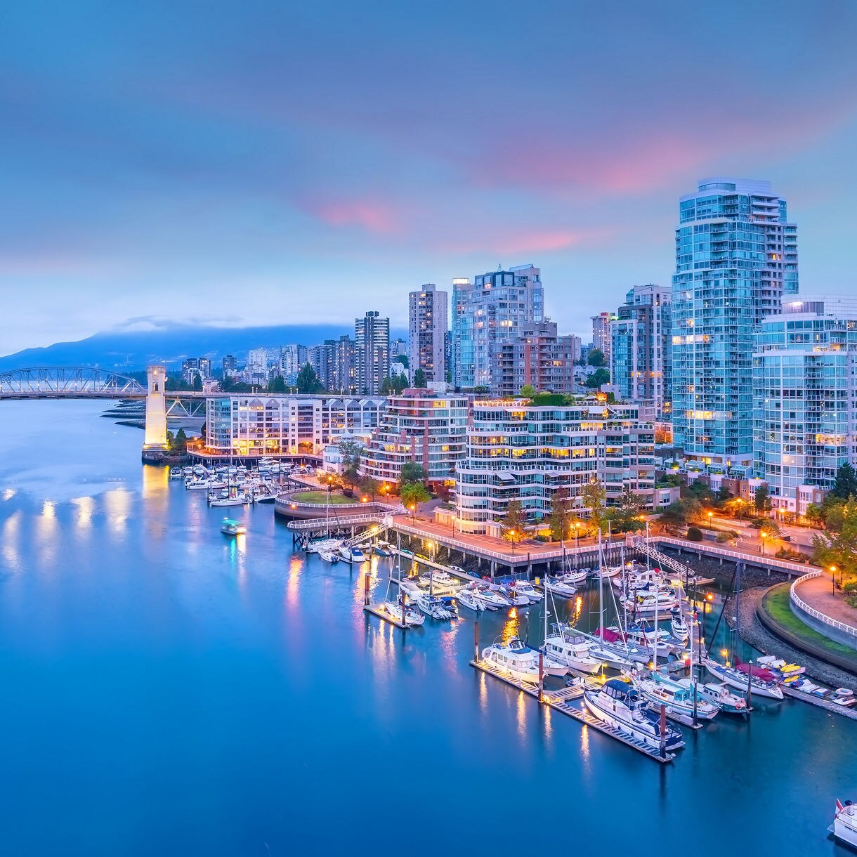 A nighttime view of the Vancouver, Canada city skyline, with a brightly lit bridge and boats in the water, and a colorful sky in the background.