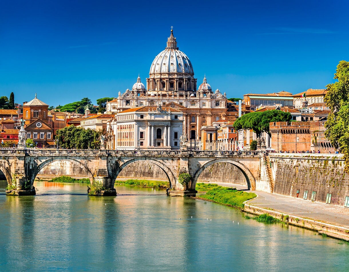 View of St. Peter’s Basilica behind the historic bridge over the Tiber River, with warm-toned buildings and bright blue sky.