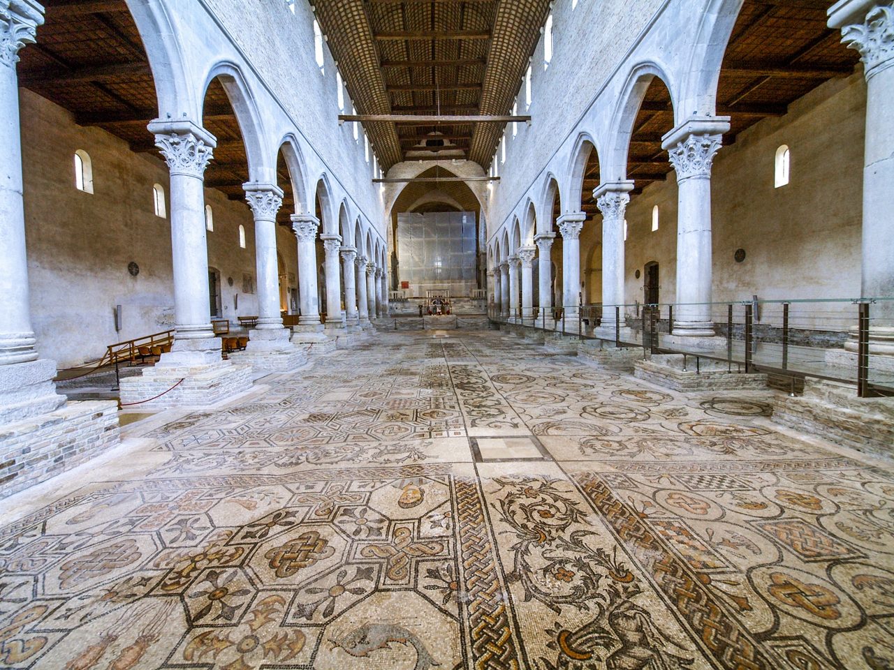  Interior of the Basilica of Aquileia in Italy, featuring tall stone columns and a vast mosaic floor decorated with intricate geometric and floral patterns.