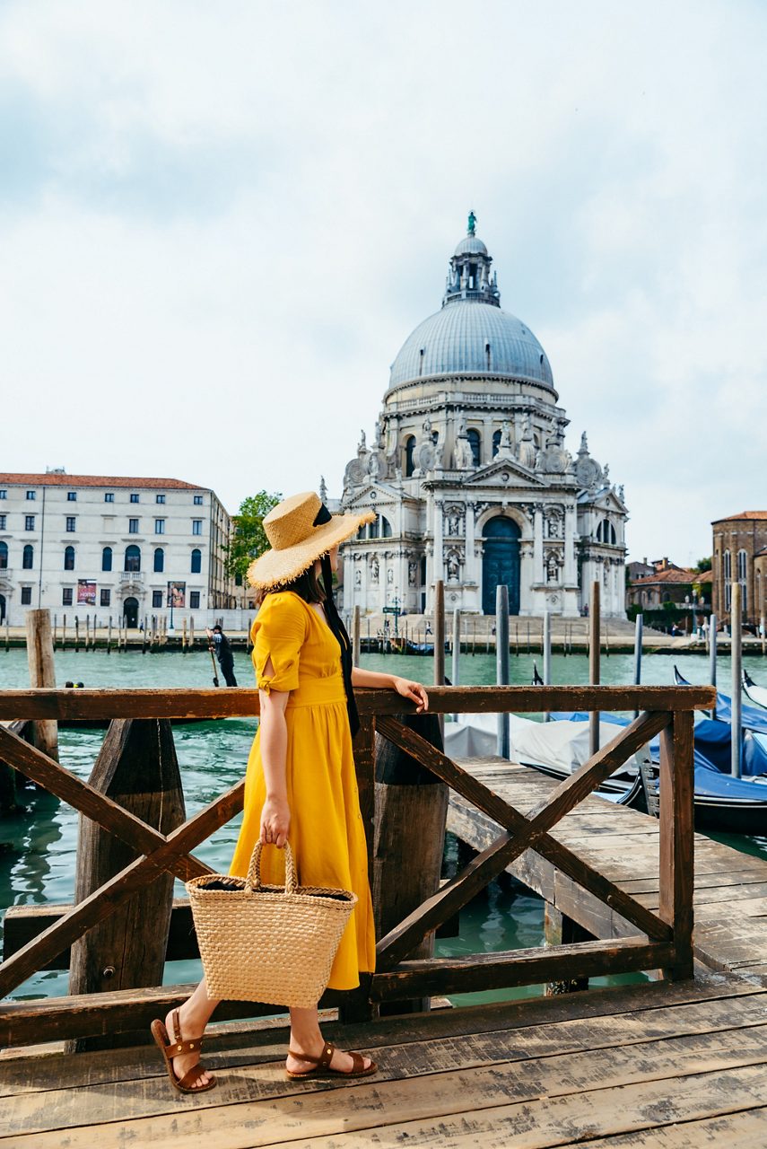  Woman in a yellow dress and straw hat standing on a wooden dock with Santa Maria della Salute in the background.