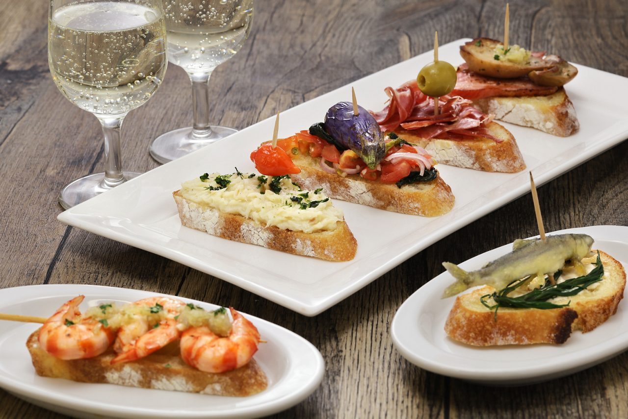 Plates of Venetian cicchetti topped with seafood, meats and vegetables beside glasses of white wine.