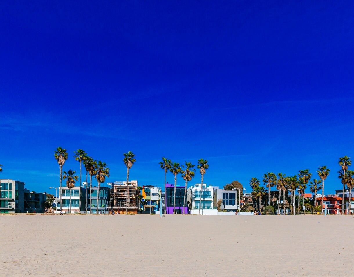 Palm trees and colorful modern homes bordering a wide sandy beach under a deep blue sky at Venice Beach.